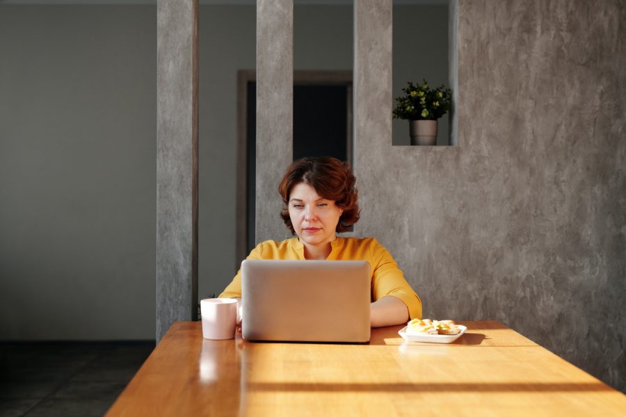 Photograph of staff member wearing a yellow sweater working on an Apple Mac laptop at a wooden table while engaging in cybersecurity when working from home.
