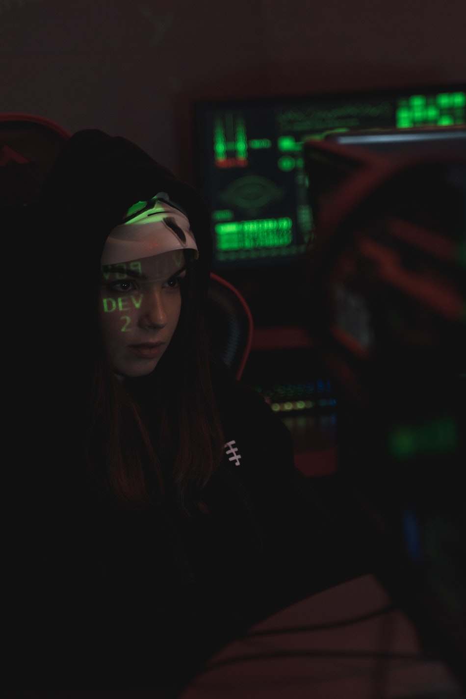 Image of a hooded foreign hacker sitting in a dark room with the computer screen shining a green shadowy glow on her face to foreshadow cybersecurity enforcement
