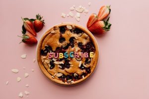 A pie surrounded by strawberries and almonds with letters spelling out subscribe dotted on top and a pink background