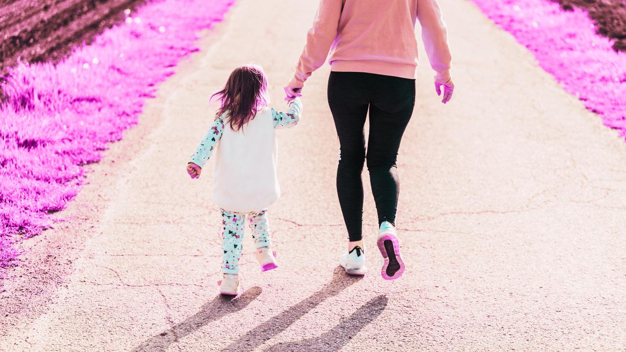 Child walking away from the camera holding hands with an adult. They're walking on a footpath with pink grass around it. The pink grass is due to a filter showing the Privacy 108 brand colours.